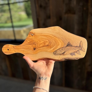 May include: A wooden cutting board with a burnt-in design of a trout fish. The board has a handle and is made of olive wood.