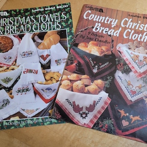 May include: Two Leisure Arts craft booklets titled Christmas Towels & Bread Cloths and Country Christmas Bread Cloths. The covers feature photographs of embroidered white and colored fabric cloths used to serve various baked goods.