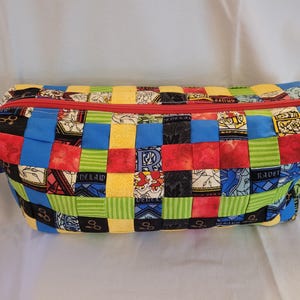 Puede incluir: Una bolsa rectangular de tela acolchada con cremallera roja. La bolsa está hecha de cuadrados tejidos en azul, rojo, amarillo, verde y negro, con varios patrones y texto. La bolsa tiene una lengüeta de tela azul en un lado.