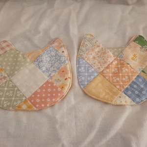 Puede incluir: Dos posavasos acolchados con forma de gato. Cada posavasos está hecho de tela de retazos en colores pastel, incluyendo rosa, azul, amarillo y verde. Los posavasos tienen forma redondeada con orejas puntiagudas.