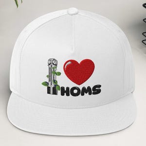 Puede incluir: Gorra de béisbol blanca con visera plana. Presenta un diseño bordado con un corazón rojo, unas tijeras y el texto "I HOMS" en negro. Un accesorio de moda.