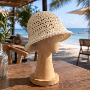 May include: A cream-colored, crocheted bucket hat with a wide brim. The hat is displayed on a wooden mannequin head. The hat has a perforated design, allowing for ventilation. The background shows a beach scene with palm trees and the ocean.