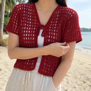 May include: A deep red, short-sleeved crochet cardigan featuring an open-knit pattern and a front tie closure. The garment is worn over a plain white top and paired with light-colored trousers that have a drawstring waist detail.