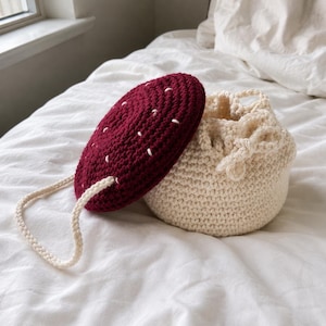 Op de afbeelding: Een gehaakte tas met trekkoord, voorzien van een crèmekleurige basis en een afneembaar, rond, donkerrood deksel met witte stikselaccenten. De tas heeft een bijpassend crèmekleurig gevlochten koord als handvat dat aan het deksel is bevestigd.