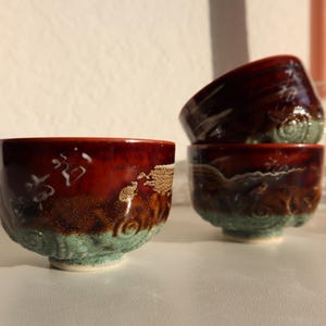 Puede incluir: Tres pequeñas tazas de té de cerámica roja y marrón con detalles blancos y dorados. Las tazas tienen un acabado texturizado y esmaltado y están apiladas una encima de la otra.