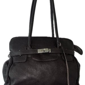 May include: A dark brown leather handbag featuring a textured surface, two long shoulder straps, and a horizontal strap across the front secured with a silver-toned metal clasp. A thin leather cord hangs from the right side of the bag.