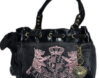 May include: Black and grey Juicy Couture handbag with a pink embroidered crest design. The bag features a drawstring closure, black handles, and a heart-shaped charm. The bag has gold-coloured hardware.
