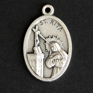 May include: Silver oval pendant with a depiction of Saint Rita of Cascia. The pendant features a halo above her head and a cross behind her. The text "ST. RITA" is inscribed around the edge of the pendant.
