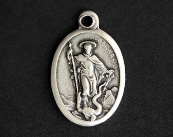 Può includere: Pendente ovale in argento con una raffigurazione di San Bernardo, che tiene un bastone e sta in piedi sopra un serpente. Il pendente ha il testo "S. BERNARD" inciso sul lato.