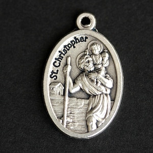 Op de afbeelding: Zilveren ovale hanger met een afbeelding van Sint Christoffel die een kind op zijn schouders draagt. De hanger heeft de woorden "St Christopher" rond de rand gegraveerd.