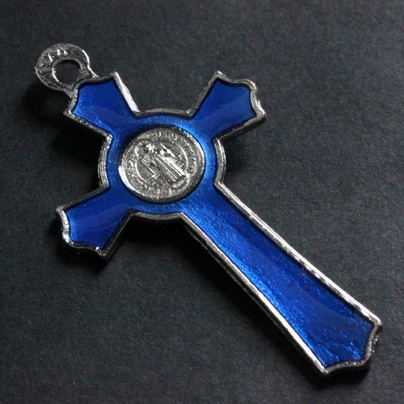 Puede incluir: Una cruz de esmalte azul con una medalla plateada que presenta una imagen religiosa. La cruz tiene un bucle en la parte superior para colgar.