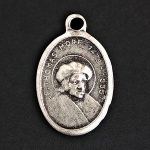 May include: Silver oval pendant featuring a portrait of Sir Thomas More, a renowned English lawyer, author, and statesman. The pendant is inscribed with "St. Thomas More 1478-1535".
