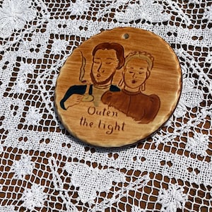 May include: A round, ceramic wall hanging with a painted illustration of a man smoking a pipe and a woman. The text "Outen the light" is below the figures. The background is a light brown color.
