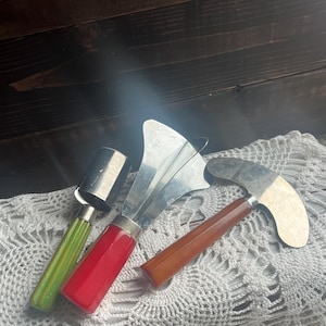 Op de afbeelding: Drie vintage keukengereedschappen met kleurrijke handvatten. Een ijslepel met groene handgreep, een appelboor met rode handgreep en een kaasmessen met bruine handgreep liggen op een wit kanten kleedje. De gereedschappen hebben zilveren metalen bladen.