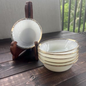 May include: A set of five white milk glass bowls featuring a swirling pattern and a gold rim. One bowl is displayed upright on a wooden stand, while the other four bowls are stacked together in a pile, showcasing the consistent design and metallic trim.