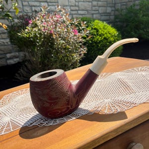 May include: A burgundy-colored tobacco pipe with a cream-colored stem and mouthpiece. The pipe's bowl features an etched design. It rests on a wooden table with a white lace doily. The background includes greenery and a stone wall.