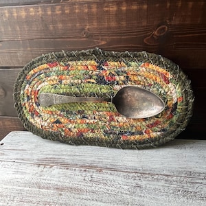 May include: An oval-shaped, braided fabric trivet with a colorful patchwork design in shades of green, orange, and red. A vintage silver spoon rests on top. The trivet is placed on a distressed white surface.