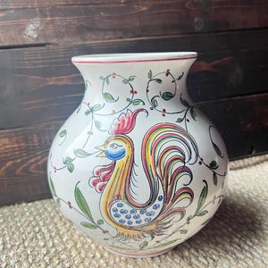 Puede incluir: Un jarrón de cerámica blanco con un diseño de gallo colorido. El gallo presenta detalles amarillos, rojos, azules y verdes. El jarrón está decorado con hojas verdes y bayas rojas. El borde tiene un borde rojo.