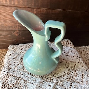 May include: A vintage turquoise ceramic pitcher with a unique, curved handle and spout. The interior of the spout has a pink hue. The pitcher sits on a white lace doily, with a dark wood background.