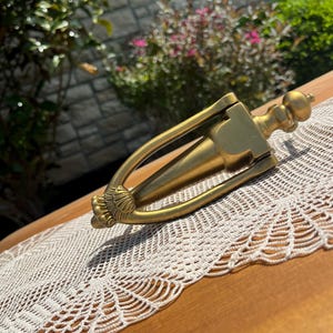 May include: A brass-coloured door knocker with an ornate design. It rests on a wooden surface, with a cream lace doily beneath. The background includes foliage and a stone wall.