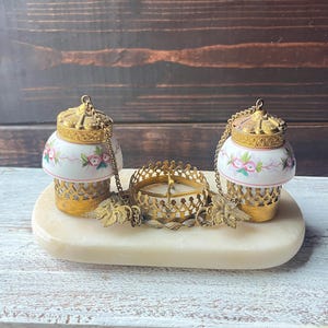 Puede incluir: Un conjunto decorativo con dos pequeños tarros con tapa, diseños florales y detalles metálicos dorados. Los tarros están conectados a un candelabro central por cadenas y se asientan sobre una base ovalada de color crema. El conjunto tiene una estética vintage.