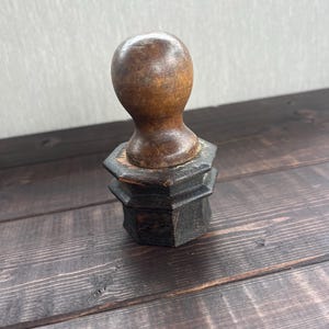 Könnte beinhalten: Ein Handstempel aus Holz mit einem glatten, abgerundeten, hellbraunen Griff, der auf einem dunklen, achteckigen Sockel befestigt ist. Der Sockel hat ein gestuftes Design mit sichtbarer Holzmaserung und Gebrauchsspuren, während der Griff poliert ist.