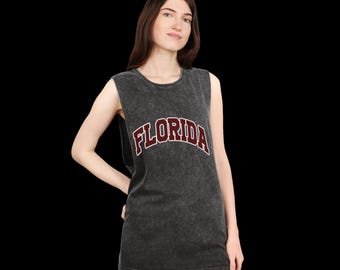 Puede incluir: Una camiseta sin mangas gris oscuro con la palabra "FLORIDA" en letras granate y blancas. La camiseta tiene un aspecto ligeramente descolorido y vintage. La persona lleva vaqueros azules.