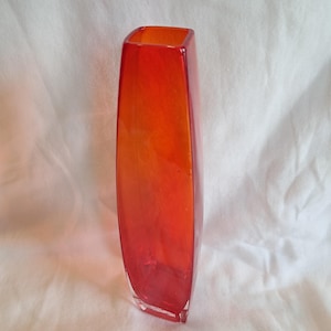 Pode incluir: Um vaso retangular alto com um gradiente de tons laranja e vermelho. O vaso de vidro tem uma abertura quadrada e uma forma ligeiramente cónica. O vaso é adequado para exibir flores ou como um elemento decorativo.