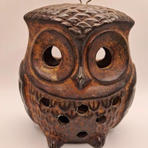 May include: A brown ceramic owl-shaped lantern featuring a textured feather pattern, large circular eye openings, and smaller circular cutouts across the chest area. A small metal loop is attached to the top of the head for hanging purposes.