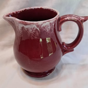 May include: A deep burgundy ceramic pitcher featuring a rounded body, a curved handle, and a distinct pouring spout. The upper rim and the top of the handle are accented with a textured, dripped white glaze that contrasts against the glossy finish.