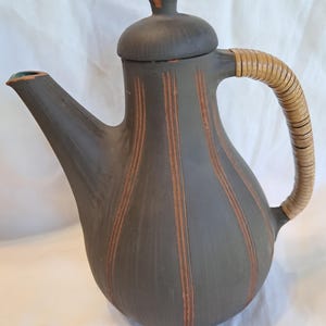 Puede incluir: Una tetera de cerámica gris oscuro con tapa y asa trenzada. La tetera tiene rayas verticales marrones y un pico con interior verde. El diseño mezcla estilos modernos y rústicos.
