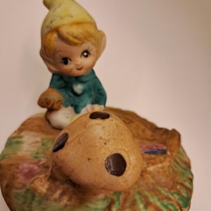 May include: A ceramic figurine of a small elf with blonde hair and a yellow hat, wearing a teal jacket. The elf sits on a textured base featuring a tan mushroom with three dark brown spots and patches of green grass and pink flowers.