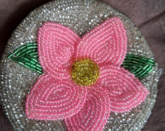 May include: A round, beaded coin purse with a silver-colored base. A large pink flower with a yellow center is beaded on the front, with green beaded leaves. The purse has a silver clasp.