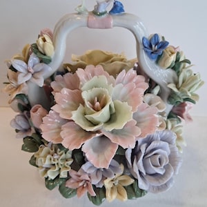 May include: A ceramic basket overflowing with pastel-colored flowers. The basket handle is white with delicate floral accents in pink, blue, and yellow. The flowers include roses, peonies, and other blooms.