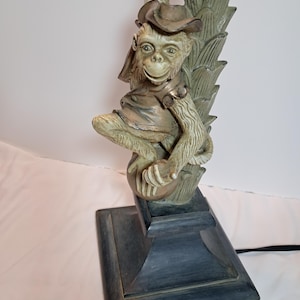 May include: A table lamp featuring a sculpted monkey wearing a hat and vest, perched on a sphere against a palm tree trunk. The monkey and tree are finished in muted green and bronze tones, set upon a square, dark-colored pedestal base.