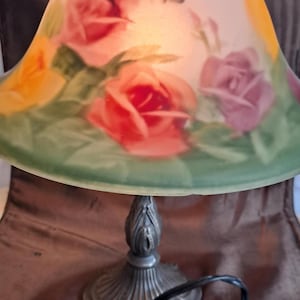 May include: A decorative table lamp with a floral design. The lampshade features red, pink, and yellow roses with a butterfly on a cream-colored background. The base is a detailed, ornate metal design with a black cord.