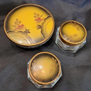 May include: Three glass vanity jars with lids featuring a yellow and black gradient finish. The lids are decorated with gold-toned floral patterns and scalloped edges. The jars have clear, faceted glass bases with a multi-sided, hexagonal shape.