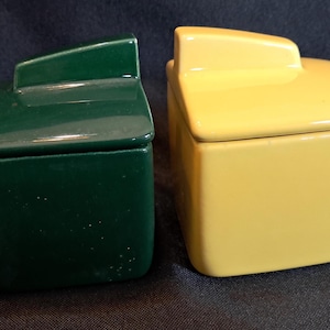 May include: Two square ceramic containers with lids, one in a deep forest green and the other in a bright yellow. Each container features a smooth, glossy finish and a distinct, raised geometric handle on the top of the lid, creating a uniform shape.