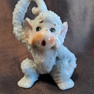 May include: A quirky, off-white figurine of a creature with a surprised expression. The figurine has a textured, fuzzy body, pink ears, and a peach-colored nose. Its eyes are detailed with black and green accents, and its mouth is open.