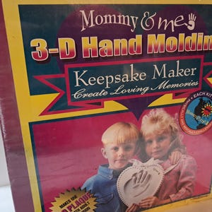 May include: A boxed 3-D Hand Molding Keepsake Maker kit. The box features the text "Mommy & Me" and "Create Loving Memories." The image shows two children holding a heart-shaped handprint plaque. The box is yellow, red, and blue.