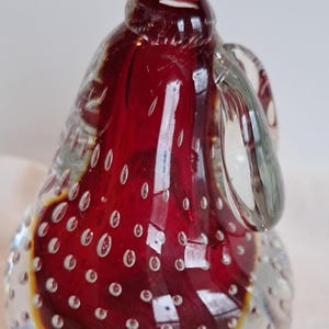 May include: A decorative glass pear sculpture. The pear is primarily a deep red color with a clear glass stem and handle. The body of the pear is filled with small air bubbles, and the base has a yellow hue.