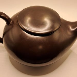 Puede incluir: Una tetera marrón con un cuerpo redondeado, un asa curva y un pico. La tapa es de color marrón a juego y se asienta sobre la tetera. La tetera está hecha de un material liso y tiene un diseño simple y elegante.