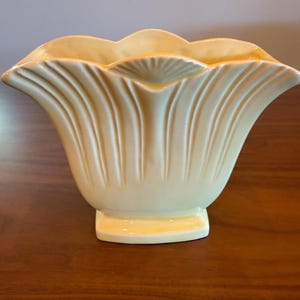 Può includere: Un vaso in ceramica giallo pallido con bordo smerlato e dettagli a coste verticali. Il vaso ha una forma svasata e poggia su una base rettangolare. Il design ricorda un fiore.