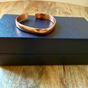May include: A copper-colored cuff bracelet with a slightly curved design. The bracelet is resting on a dark blue box, set against a wooden surface. The bracelet has a smooth, polished finish.