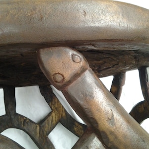 Pode incluir: Close-up de um banco de madeira artesanal. O banco apresenta um assento redondo e pernas e suportes intrincadamente esculpidos. A madeira tem uma cor castanha escura rica, com grão e textura visíveis. O design inclui padrões geométricos e elementos curvos.
