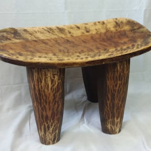 May include: A handcrafted wooden stool with a rectangular seat and three sturdy legs. The seat features a natural wood grain pattern with shades of brown and tan. The legs have a textured, bark-like appearance, adding to the rustic aesthetic.