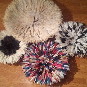 May include: Five decorative Juju hats with a circular design. The hats are made of feathers in various colors, including cream, black, and a vibrant mix of red, blue, and yellow. They are arranged on a wooden surface.