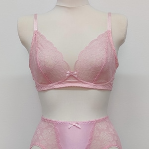 May include: A light pink lingerie set displayed on a mannequin. The set includes a lace-detailed bra with a small bow and matching garter belt with suspender straps. The bra and garter belt are made of a soft, delicate fabric.