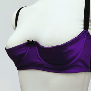 May include: A purple bra with black trim and straps. The bra has a satin-like finish and a small black bow at the center. The bra is displayed on a white mannequin.