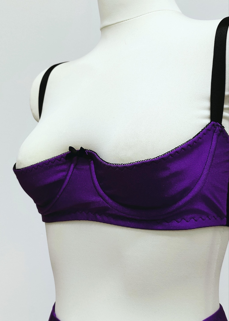 May include: A purple bra with black trim and straps. The bra has a satin-like finish and a small black bow at the center. The bra is displayed on a white mannequin.